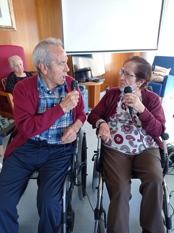 Dos residentes sentados en sillas de ruedas, cada uno sosteniendo un micrófono y cantando mientras se miran