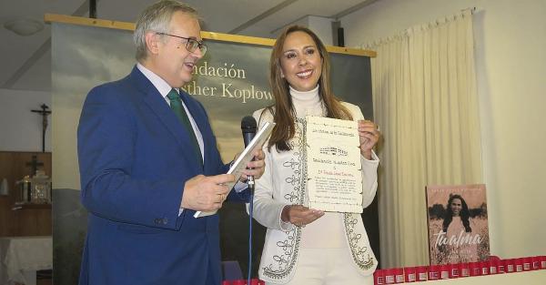 Paula López Espinosa sosteniendo un certificado de gratitud otorgado por la residencia Nuestra Casa. Junto a ella, sobre una mesa, se encuentra su libro, cuya portada incluye una fotografía suya