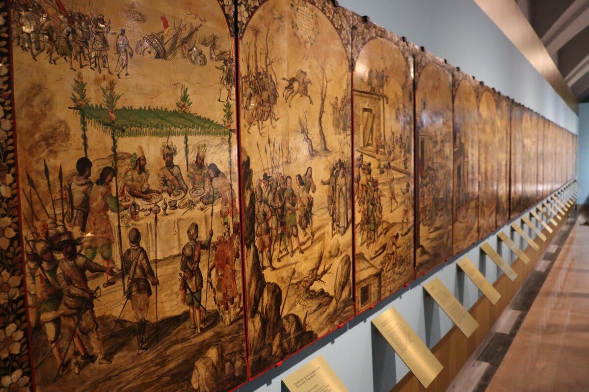 Exposición en el Museo de América con varias pinturas dispuestas horizontalmente, mostrando escenas con soldados y armas