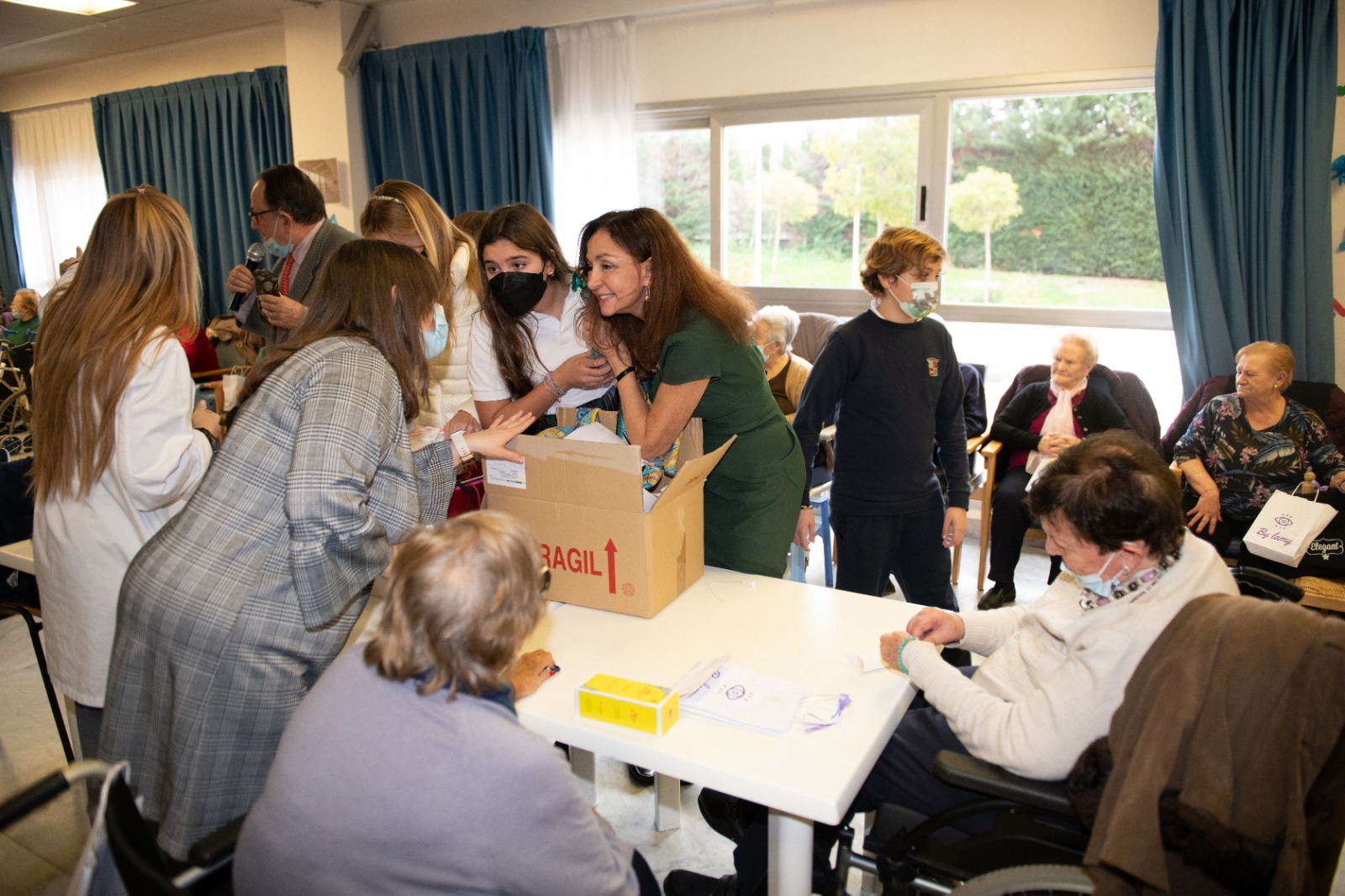 Esther Koplowitz, sonriente junto a su equipo frente a una mesa con una gran caja de regalos para los residentes; al fondo, residentes sentados en fila