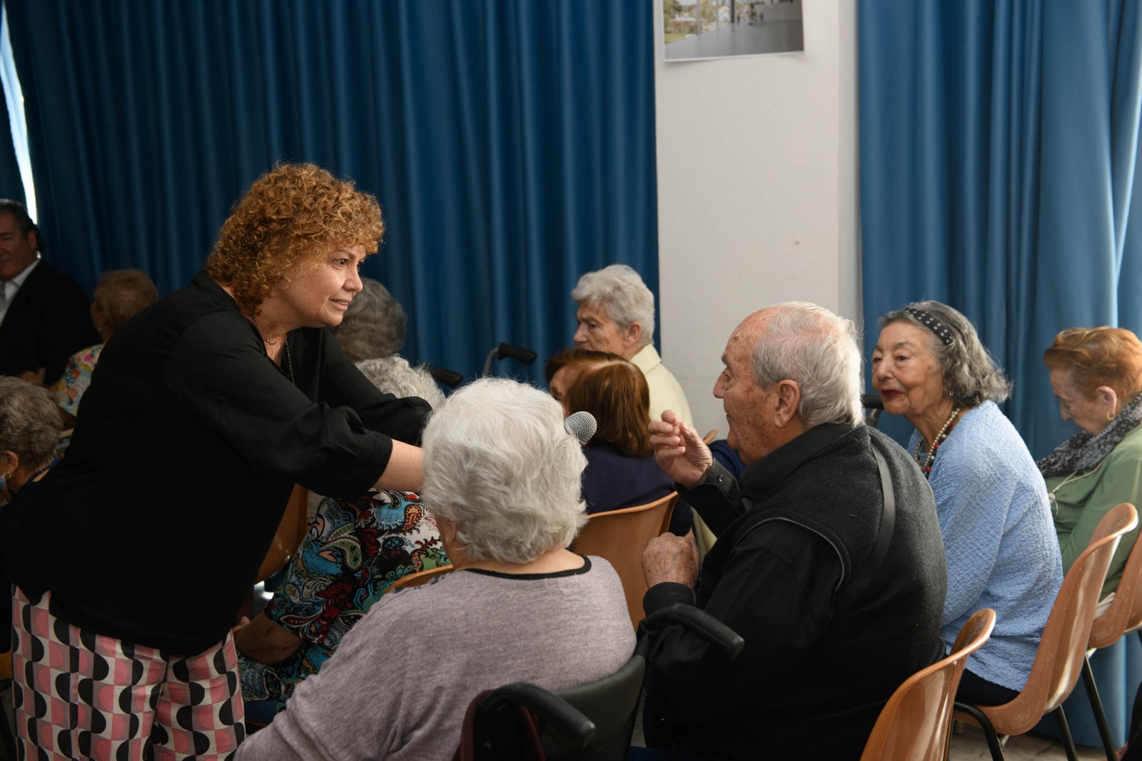 Emilia Adán García entrega el micrófono a un señor mayor durante su charla, animándolo a compartir sus propias experiencias mientras los demás asistentes observan con interés