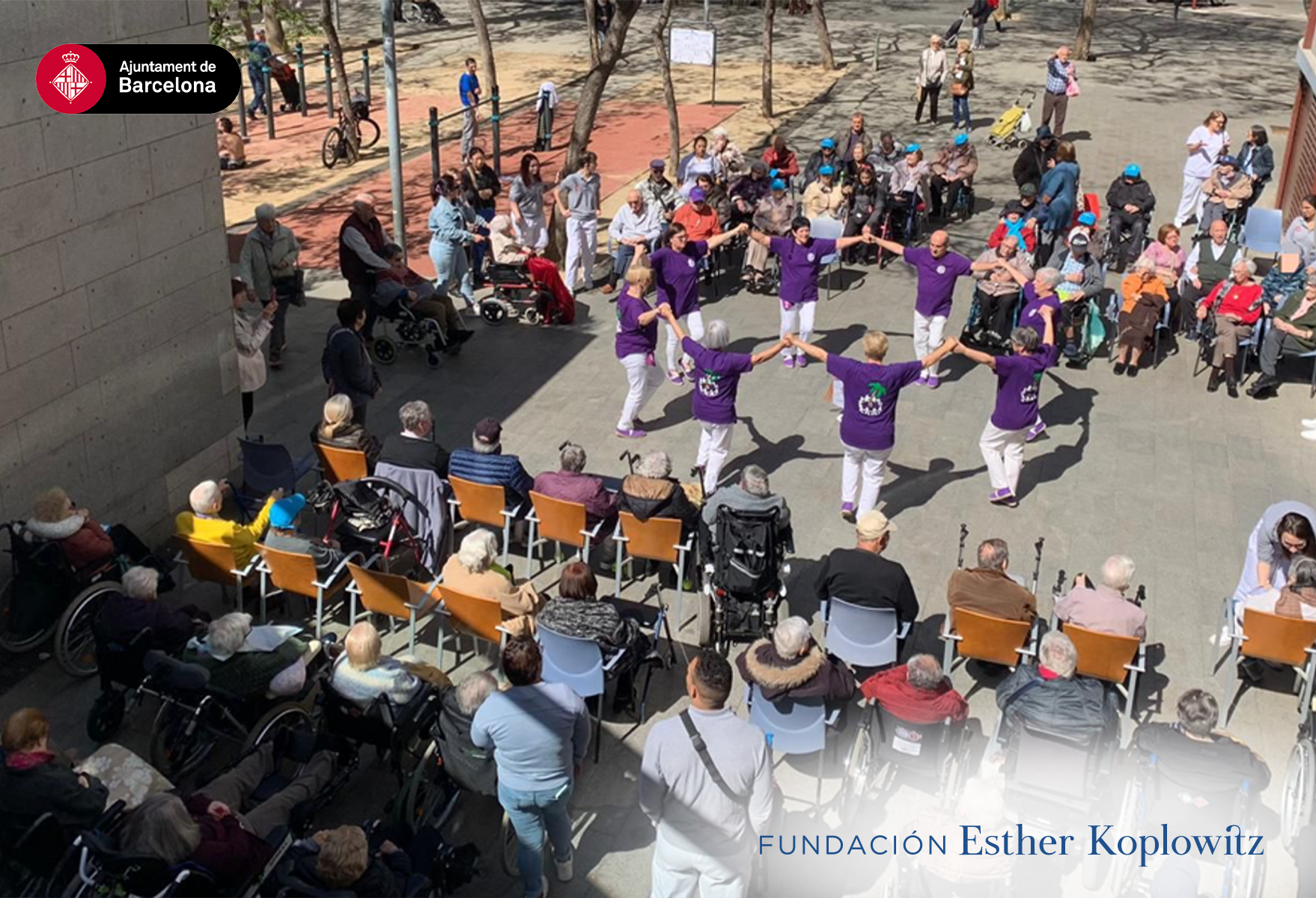 Grupo de mayores formando un círculo y cogiendo las manos, bailando juntos, mientras otros mayores están sentados disfrutando al verlos